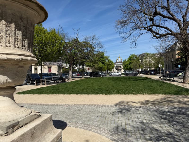 A large grassy area with trees and a paved walkway, surrounded by parked cars and buildings.