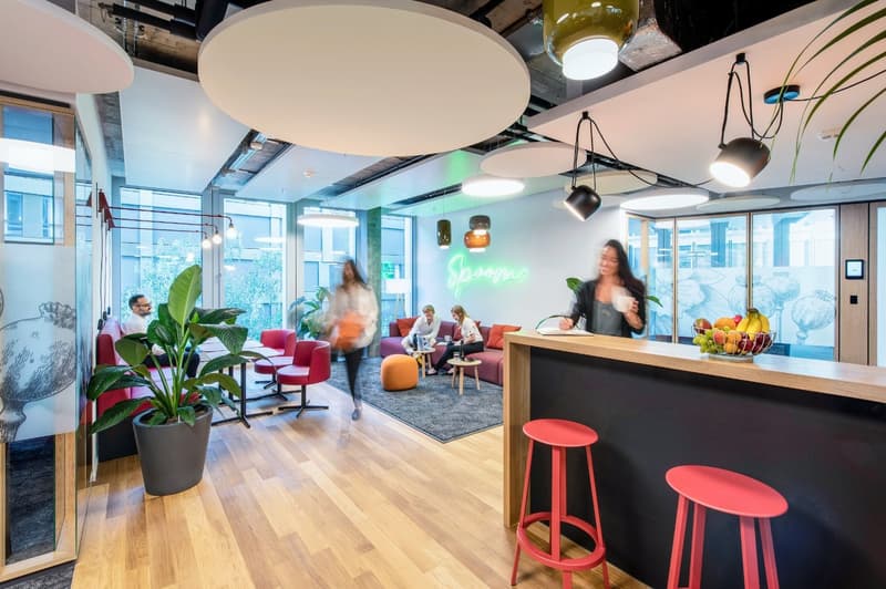 Open concept office space with large windows, modern furniture, plants, and a neon sign. The space has a kitchen area with a dishwasher and bar stools. People are working and relaxing in the different seating areas.