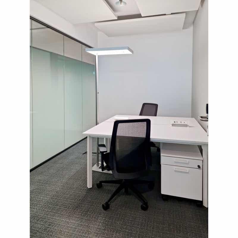 The image shows a modern office space with a white desk, a black office chair, and a glass partition wall. The room has a clean and minimalist design, with a ceiling light fixture and carpeted flooring. There appears to be a dishwasher visible in the room