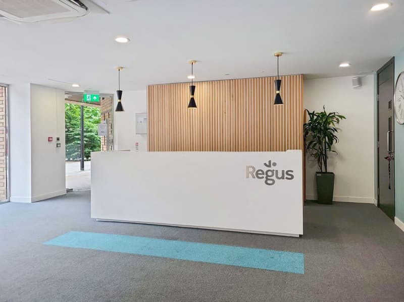 The image shows the lobby or reception area of an office space. The space features a large white reception desk with the Regus logo, wooden slat wall paneling, black pendant lights, and a potted plant. The floor is covered in a gray carpet with a teal acc