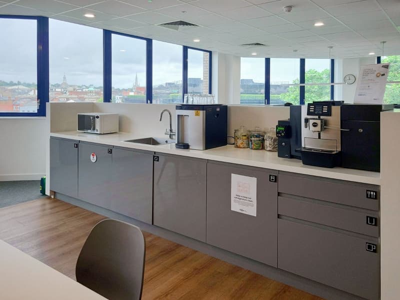 The image shows a modern office kitchen area with a sink, microwave, coffee machine, and other kitchen appliances. The kitchen has gray cabinets and countertops, and large windows overlooking the city skyline. The space appears to be designed for employee