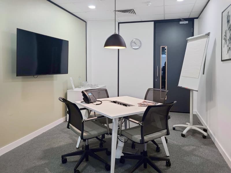 The image shows a modern office space with a large TV screen, a conference table with chairs, a whiteboard, and various office equipment such as a phone and a printer. The room has a minimalist and professional design with white walls, gray carpet, and bl