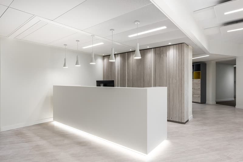 Large open office space with a white reception desk, pendant lighting fixtures, and a textured wall panel feature. The space has a modern, minimalist design with a clean and bright atmosphere.