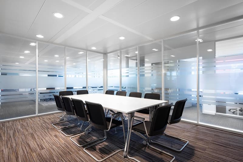 Large conference room with a long white table, black leather chairs, and floor-to-ceiling glass walls providing a view of the surrounding office space. The room is well-lit with recessed lighting in the ceiling. The floor is covered in a striped carpet pa