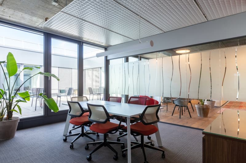 Modern office space with large windows, open floor plan, conference table, red office chairs, and potted plants.
