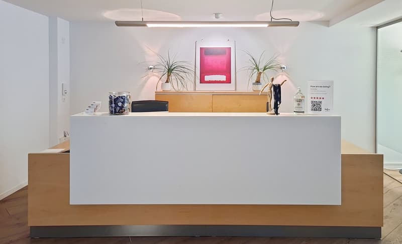 white reception desk, wooden desk, 2 potted plants, picture, glass cabinet, counter, chair, floor mounted lamps, light switches, QR code sign