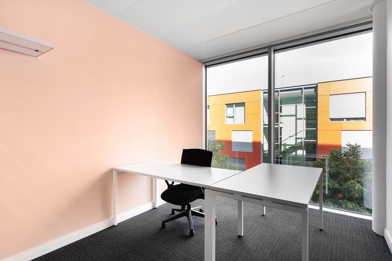 Spacious office with large windows providing a view, white desk, black office chair, peach-colored walls, and colorful exterior building visible through the windows.