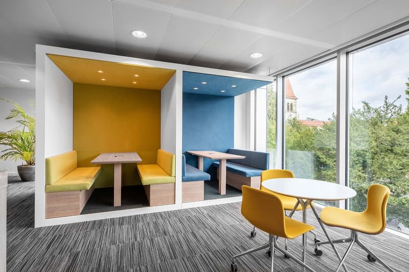 This image shows a modern and colorful office space with large windows providing a view of the surrounding greenery. The space features a yellow and blue color scheme, with yellow chairs and a yellow seating area, as well as a blue seating area. There is