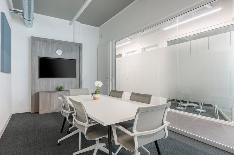 Large conference room with a white table, gray chairs, a TV mounted on the wall, and a clock. The room has large windows providing a view, and the walls are a light gray color. The floor is covered in dark gray carpet.