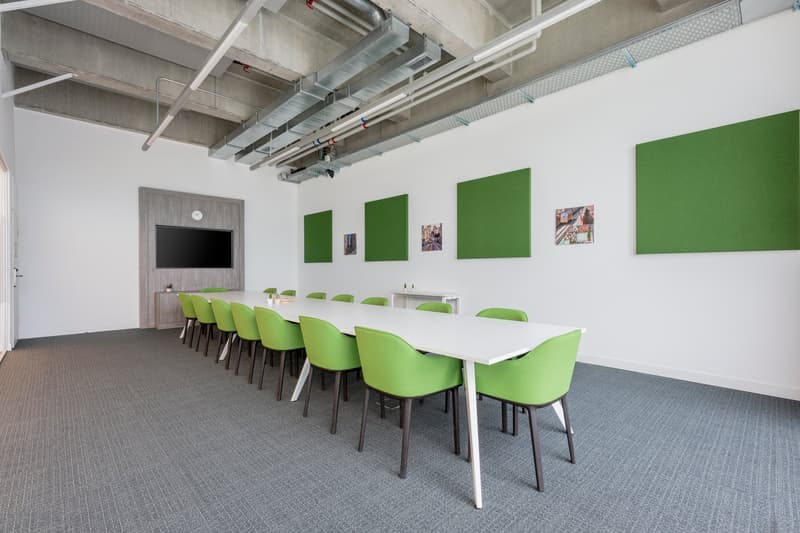 Large open meeting room with white walls, green accent panels, long white table, and green chairs. Exposed ceiling with HVAC ducts. Carpeted floor.