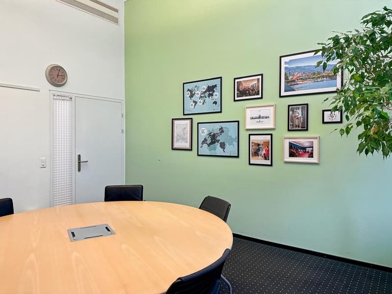The room is a conference room with a round table and four chairs. It features a green wall adorned with picture frames and a plant. There is also a clock and a door. The room has a modern design and appears to be well-lit.
