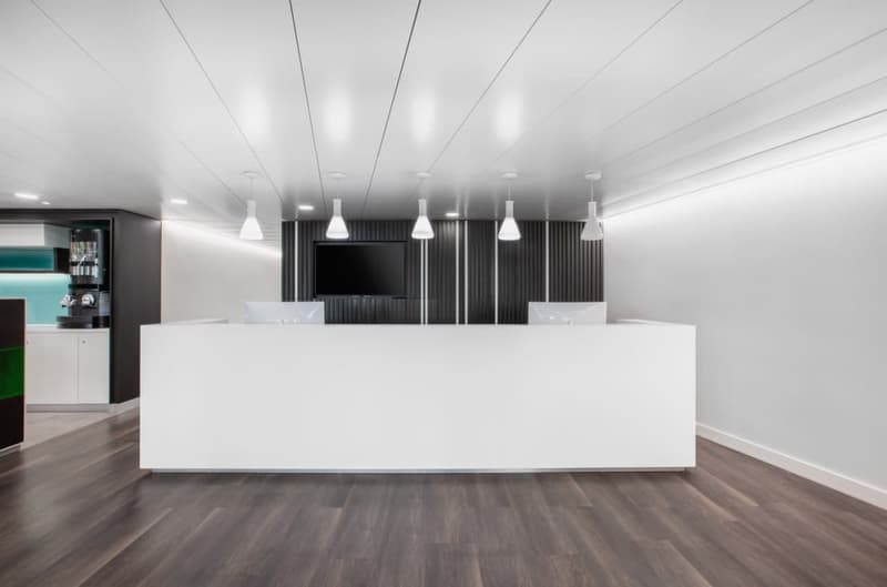 Large open office space with white reception desk, black accent walls, and multiple pendant lights hanging from the ceiling. The floor is made of dark hardwood. There is a small kitchenette area visible in the background.