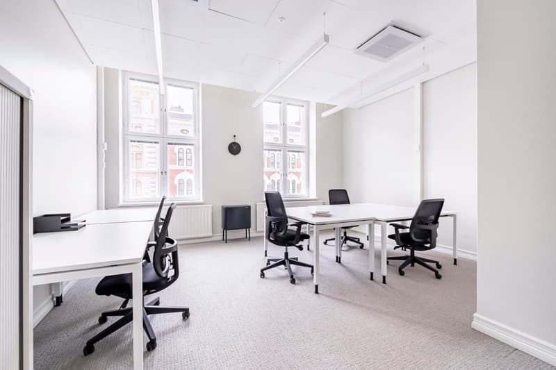 Spacious office space with large windows, white walls, and modern furniture. The room features a long table with several black office chairs, as well as individual desks and chairs. The space appears to be well-lit and designed for a professional work env