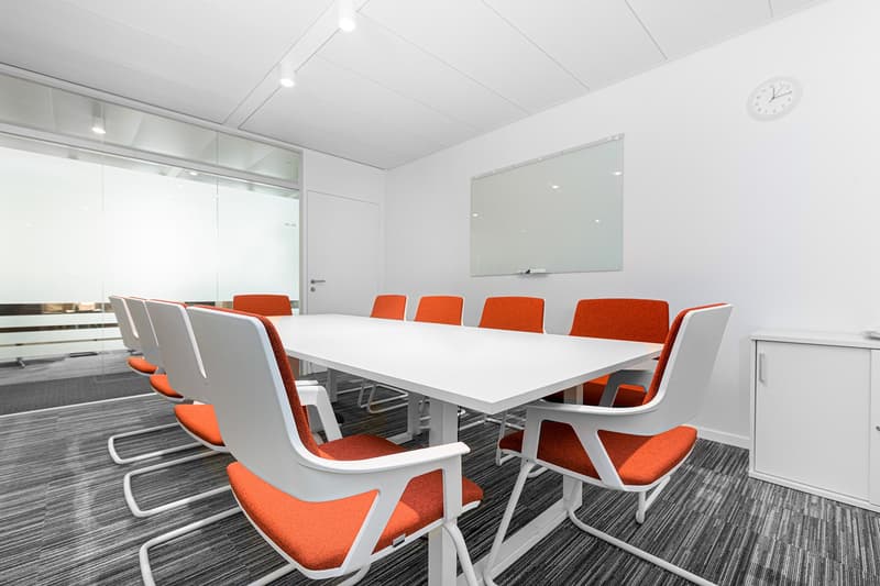 Large white conference table with orange chairs, white walls, glass windows, and a whiteboard on the wall