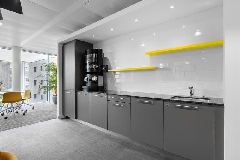 Modern kitchen with gray cabinets, black appliances including a coffee machine, yellow accent shelves, and large windows providing natural light.