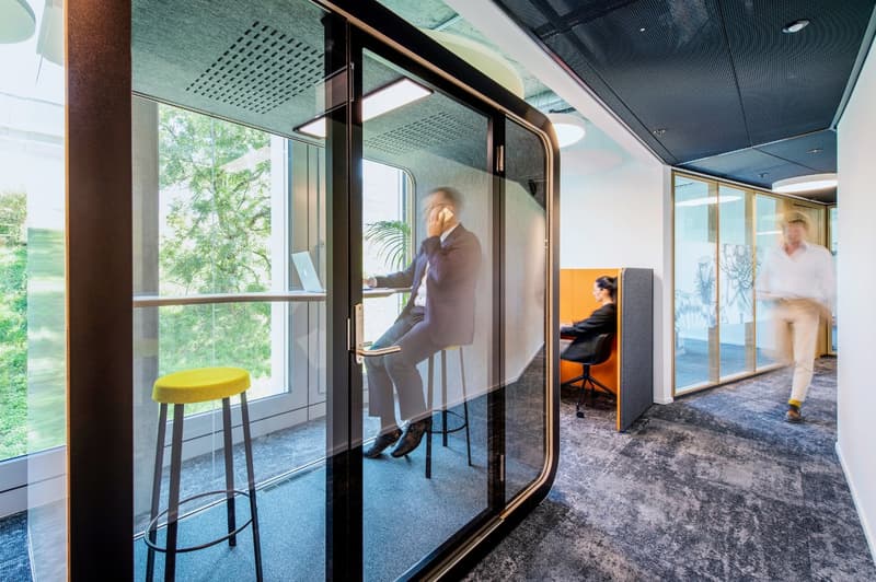 Modern office space with glass walls, high ceilings, and a view of the outdoor greenery. The space includes a bar-height table and stool, as well as private workspaces with chairs.