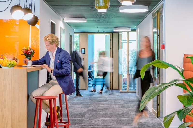 Modern and colorful office space with open layout, high ceilings, and large windows. The space features a kitchen area with a dishwasher, as well as various seating and work areas. The decor includes plants, bright lighting fixtures, and a mix of furnitur
