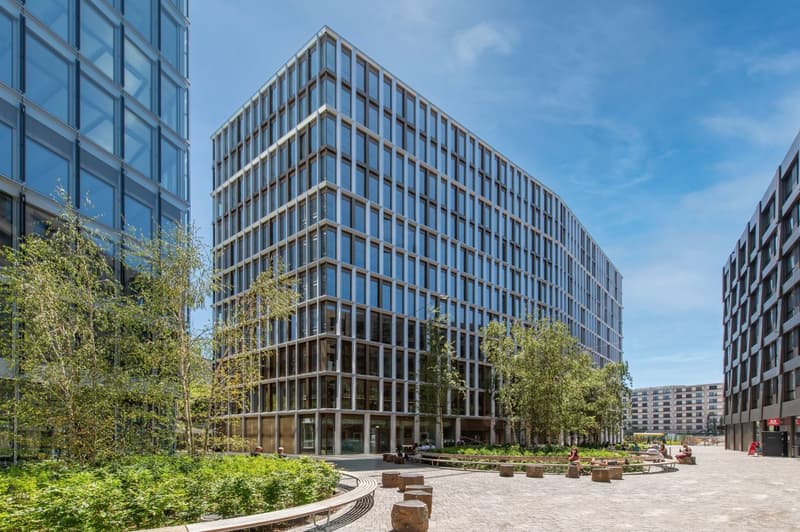 A modern, multi-story office building with a glass facade and wooden accents. The building is surrounded by lush greenery, including trees and landscaped areas. There is a paved plaza in front of the building with seating areas.