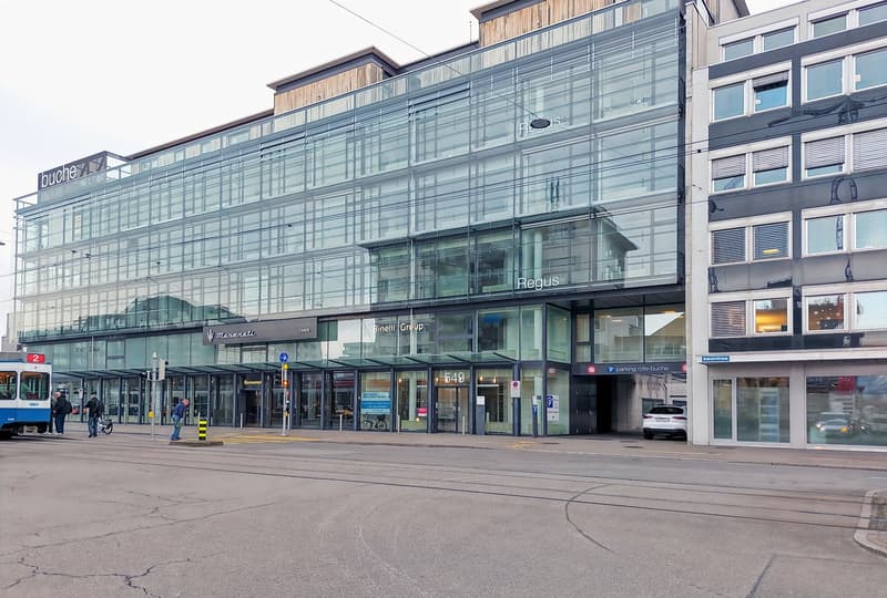A multi-story commercial building with a glass facade, housing various businesses such as Buche, Regus, and Binelli Group. The building has a parking lot in front and appears to be located in an urban area with a tram station nearby.