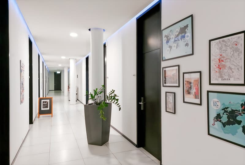 Spacious hallway with white walls, recessed lighting, and framed artwork and posters on the walls. There is a potted plant in a modern planter and a small chalkboard sign on an easel. The hallway leads to several doors and appears to be part of a larger o