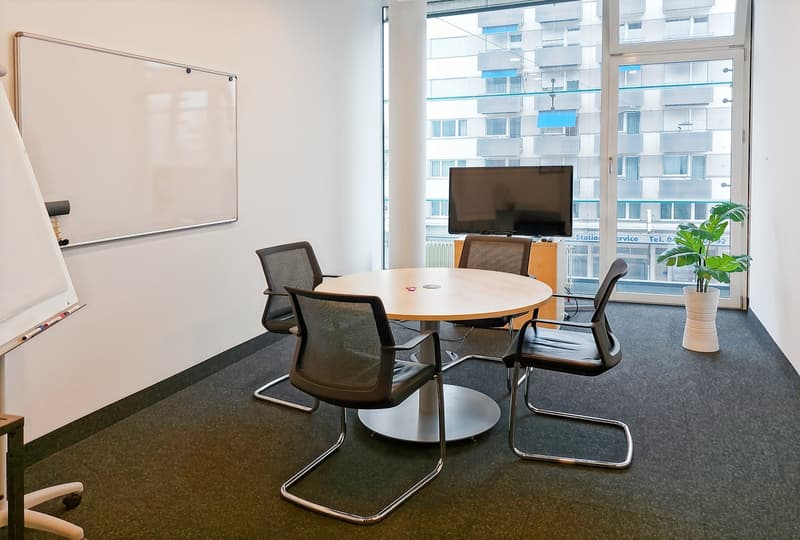 Spacious meeting room with large windows providing a view of the city, round wooden table, black mesh chairs, whiteboard, and a potted plant.