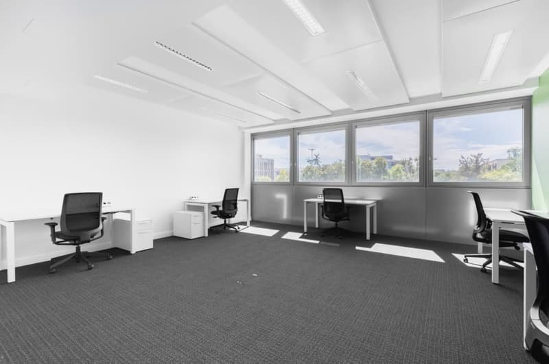 Large open office space with white walls, gray carpeted floor, multiple desks and office chairs, and large windows providing natural light and a view of the outdoors.