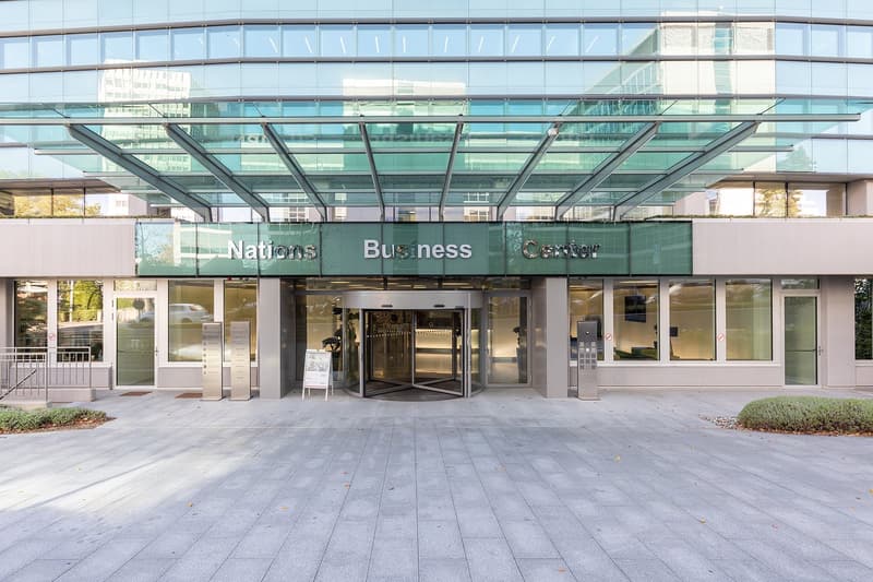 A modern glass and steel building with the signage 'Nations Business Center' displayed prominently on the facade. The entrance features a revolving glass door and there appears to be a parking area in front of the building.