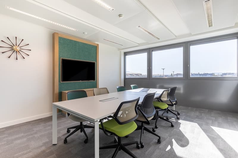 Large conference room with a long table, office chairs, a TV mounted on the wall, and large windows providing a view of the city outside.