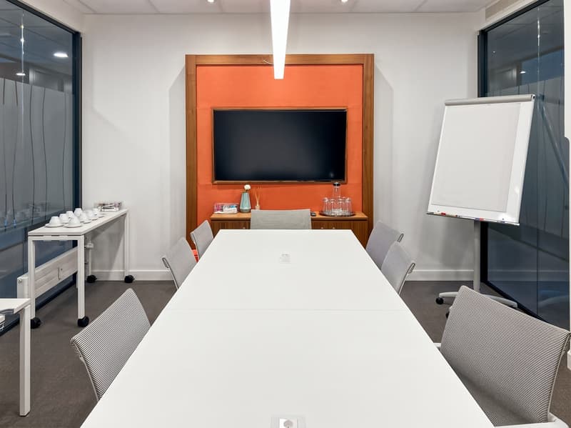 Large white conference table with chairs, TV mounted on orange wall, shelving unit, and a whiteboard on the side
