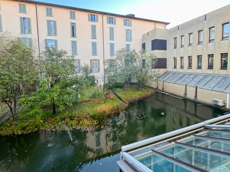 A multi-story residential building with a courtyard featuring a pond and greenery. The building has a traditional European architectural style with large windows and balconies.