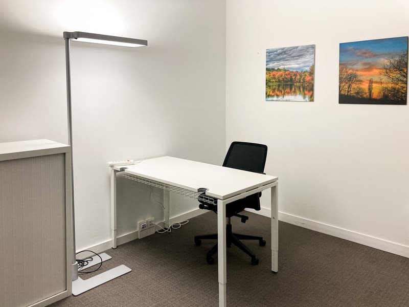 The image shows a small office space with a white desk, a black office chair, and a tall floor lamp. The walls are decorated with two landscape paintings. The room appears to be well-lit and organized for a professional work environment.