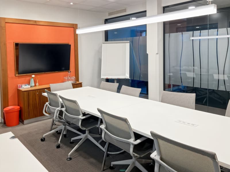 Large conference table with gray chairs, TV mounted on the wall, whiteboard, and modern lighting fixtures. The room has a sleek, professional design with a mix of wood and glass elements.
