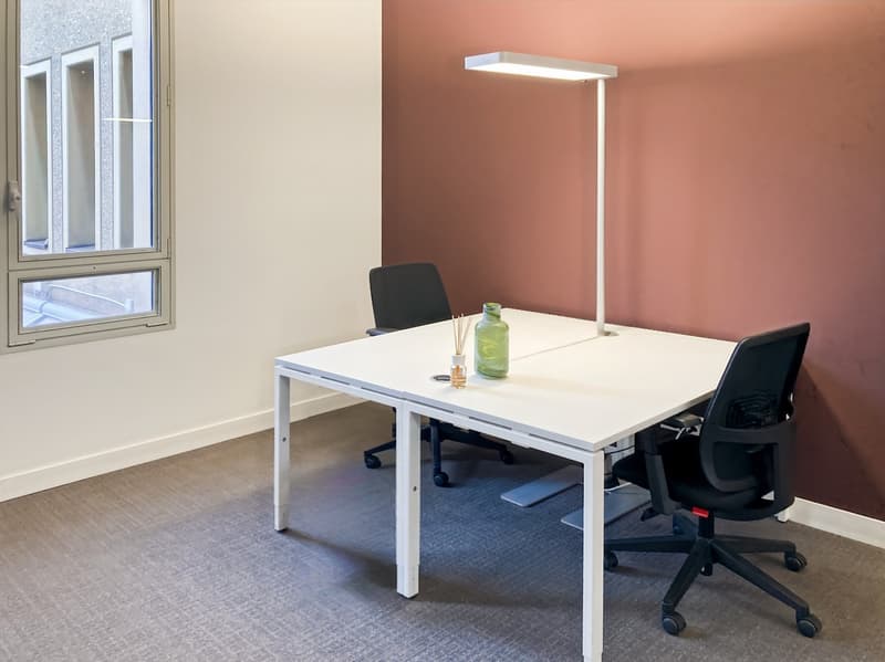 The image shows a modern office space with a white desk, a black office chair, and a floor lamp. The walls are painted in a light peach color, and the floor is covered with a gray carpet. The room has large windows that provide natural light.