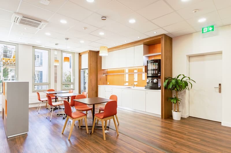 modern office breakroom with wooden floors, white walls, orange chairs, small tables, overhead lighting, plant, counter with coffee machine