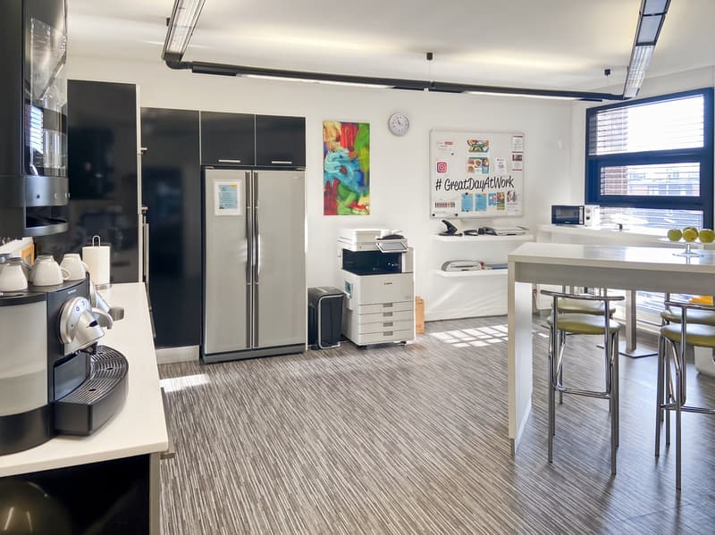 Modern office kitchen with stainless steel refrigerator, dishwasher, coffee maker, and printer. The room has white walls, a large window, and a dining table with stools. The floor is covered in a striped carpet pattern.