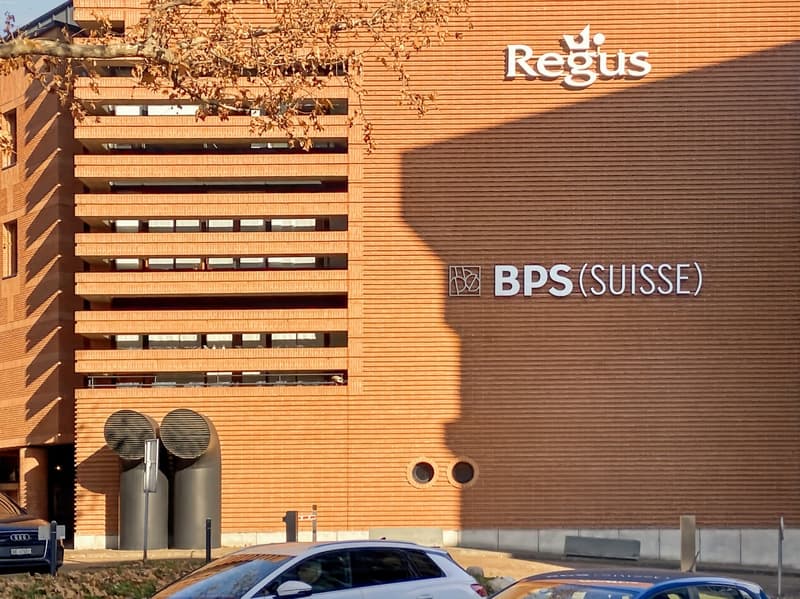 The image shows a large multi-story building with a prominent Regus logo and BPS (SUISSE) logo on the facade. The building has a modern, industrial design with a facade made of wooden panels. There are several windows visible on the upper floors of the bu