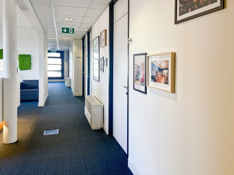 The image shows a well-lit office hallway with white walls, framed artwork on the walls, and a blue carpet floor. There is a green exit sign and a blue seating area visible. The hallway appears to be clean and well-maintained, with a professional and mode