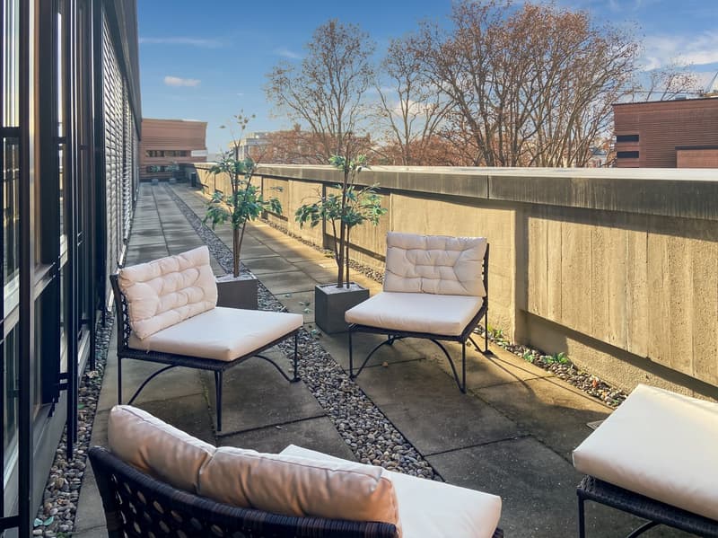 Outdoor patio area with comfortable seating, including two armchairs and a sofa. The patio has a concrete floor with gravel accents, and is surrounded by a concrete wall. There are small potted plants placed along the wall, adding some greenery to the spa