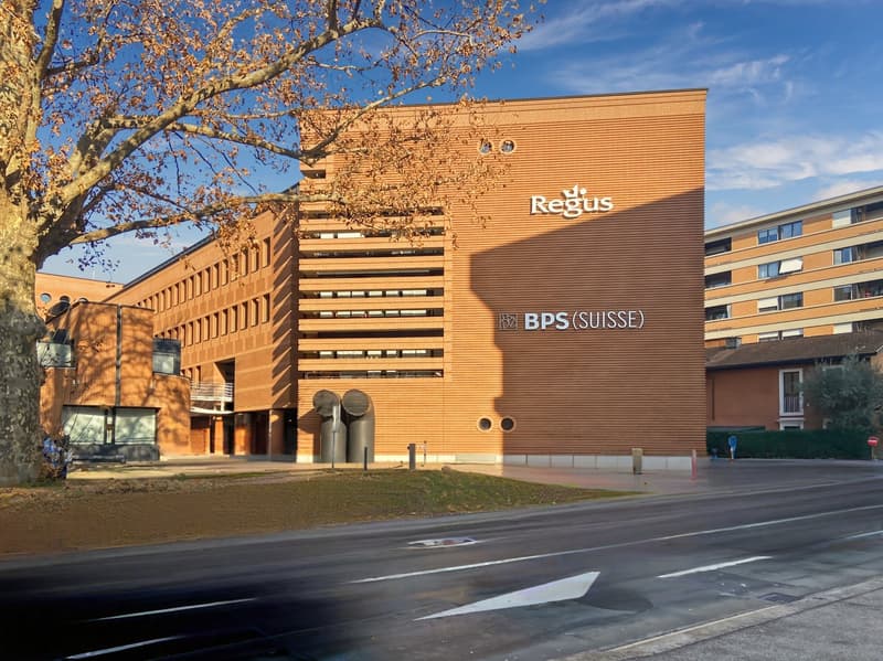 Large brown brick building with the Regus and BPS (SUISSE) logos displayed prominently. The building has a modern, striped facade design and is surrounded by trees and other buildings.