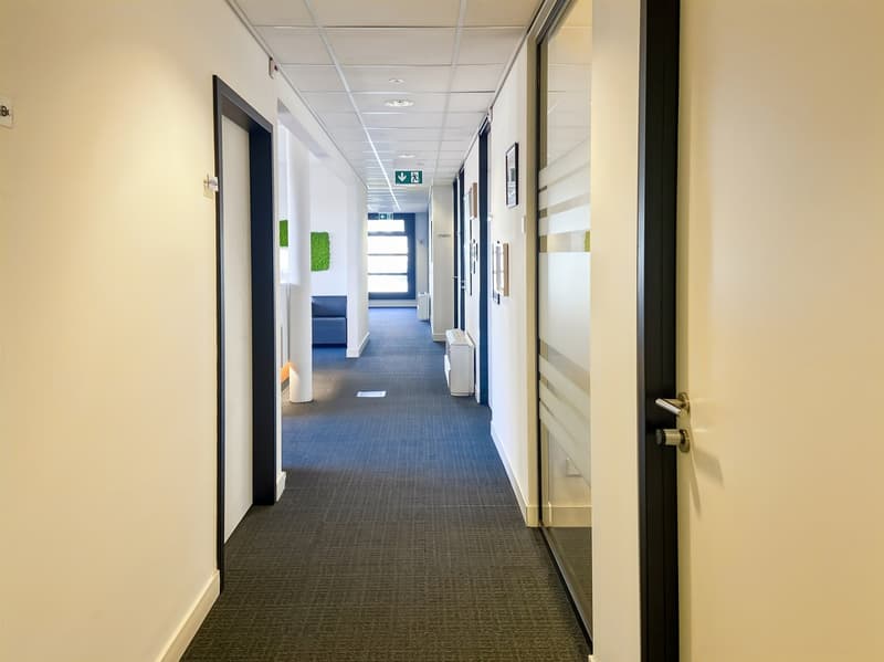 The image shows a long, well-lit office hallway with a tiled floor and suspended ceiling. The walls are painted in a light color, and there are several doors leading to different rooms or offices along the hallway. The hallway appears to be part of a larg