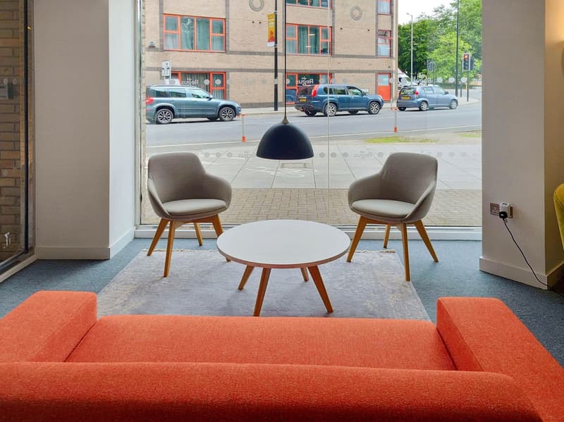 Modern furnished living room with a large glass wall, large blue sofa, two gray chairs, a round white coffee table, a hanging lamp, and visible outdoor view. The floor is covered with gray carpet.