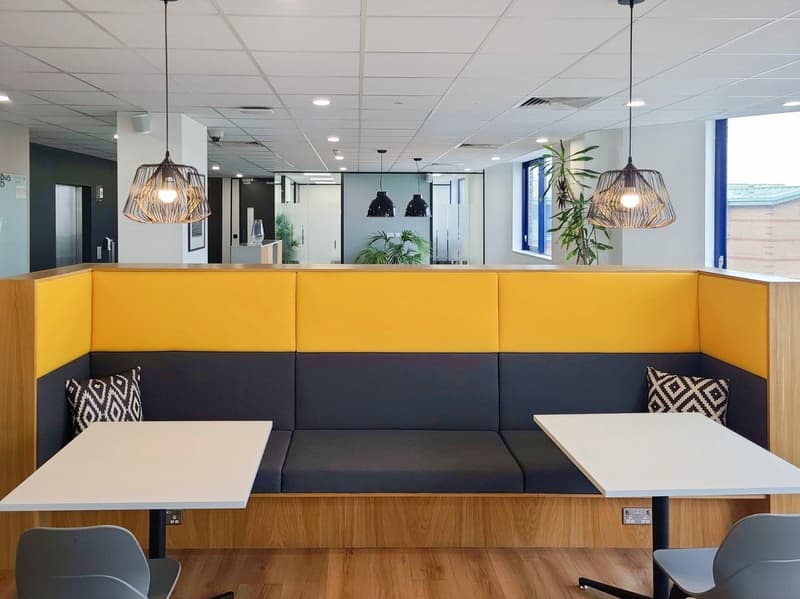 2 tables, 2 chairs, sofa, 2 cushions, pendant lights, yellow and grey partition, wooden floor, view to office spaces