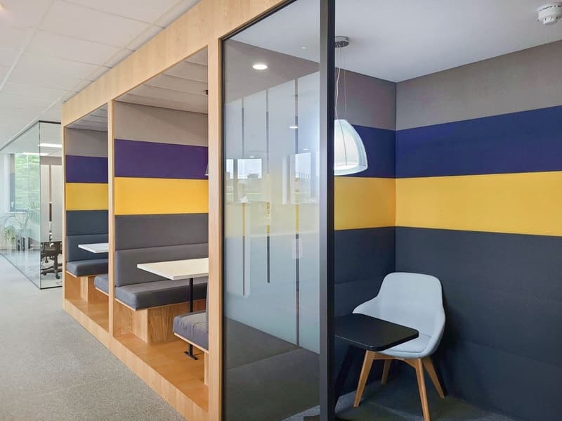 A small office space with a bench seating area, tables, and a single desk. The walls are multicolored with benches and tables matching the wall colors. There is a desk with a chair, and a lamp is hanging from the ceiling. There is a large window in the ba