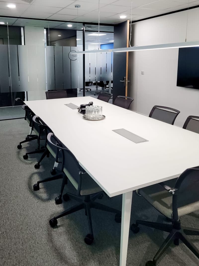Empty meeting room with a large white table, black chairs, glass dividers, fluorescent ceiling lights, a large monitor on the wall, and carpeted floor.