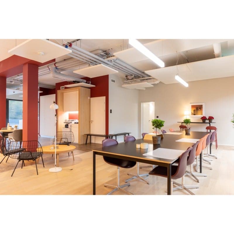 Open concept office space with hardwood floors, white walls, and exposed ceiling pipes. The space includes a large conference table with chairs, a kitchen area with a dishwasher, and various seating areas with modern furniture.