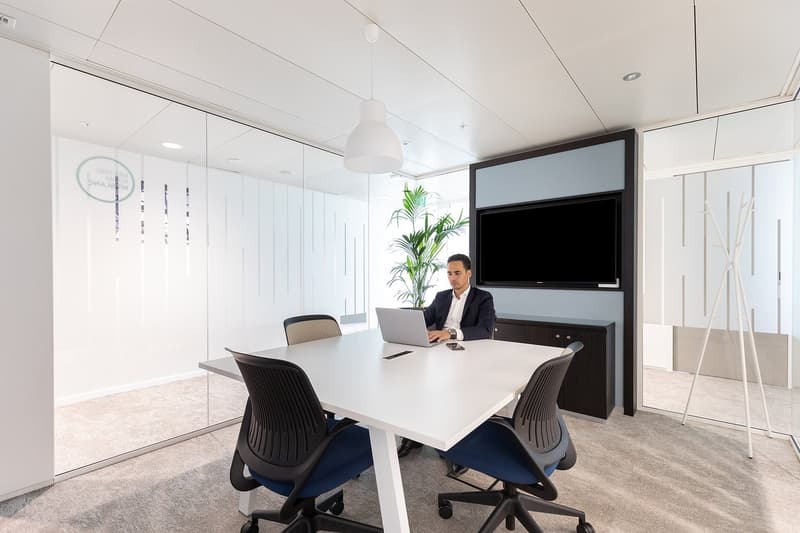A modern meeting room with a white desk, black and blue office chairs, a laptop on the desk, a person sitting, and a mounted monitor with a plant in front of the wall.