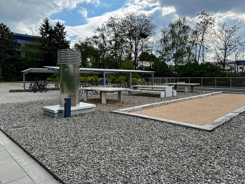 Outdoor area with gravel, picnic tables, metal structure, bicycle shed, metal structure, and a grassy area.