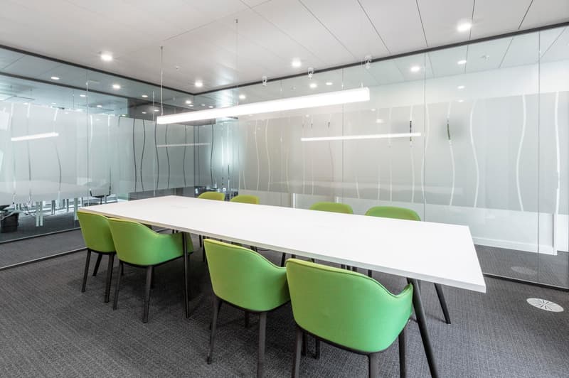 An empty meeting room with a large white table, green chairs, glass walls, gray carpet, and suspended white lights.