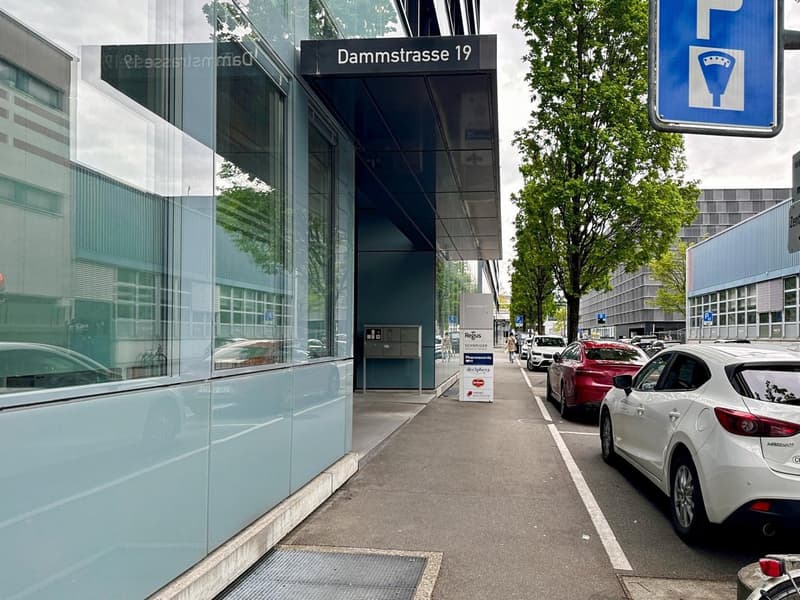The image shows a street view with a glass building on the left, a street sign indicating 'Dammstrasse 19', and several parked cars on the street. There is a parking sign visible, indicating the presence of a parking place.