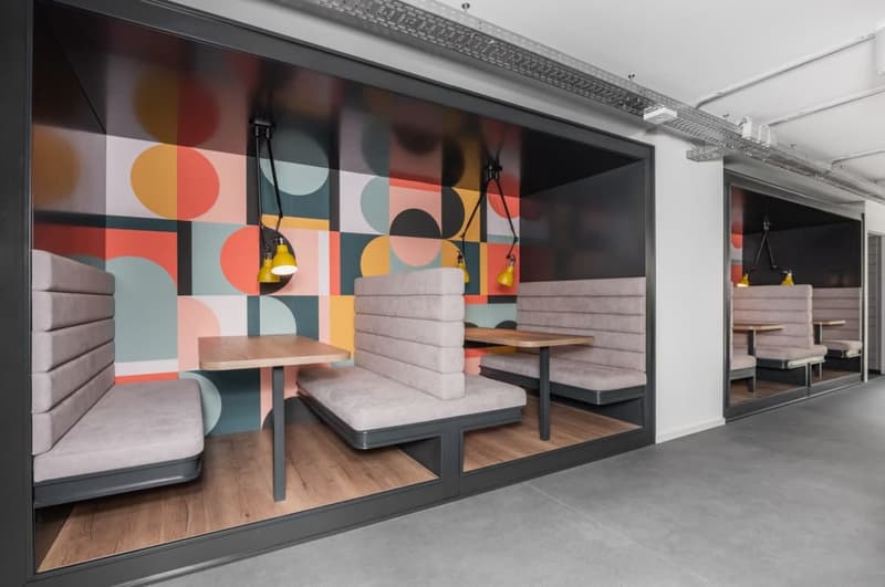 Modern and colorful office or cafe space with geometric patterned wall, wooden tables and benches, and hanging yellow lamps.